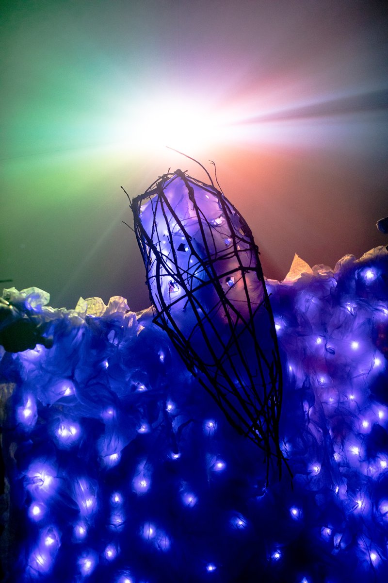 Installation view of a blue, illuminated, cocoon-like form surrounded by dark branches atop a blue, crinkled surface with embedded lights. A bright light source shines from above, casting a green and orange glow.