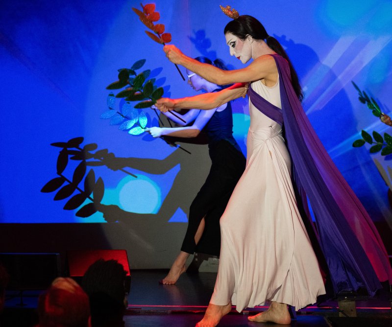 Drag queen in white gown and purple cape with crown and exaggerated makeup, holding a red leaf branch, with another performer behind.