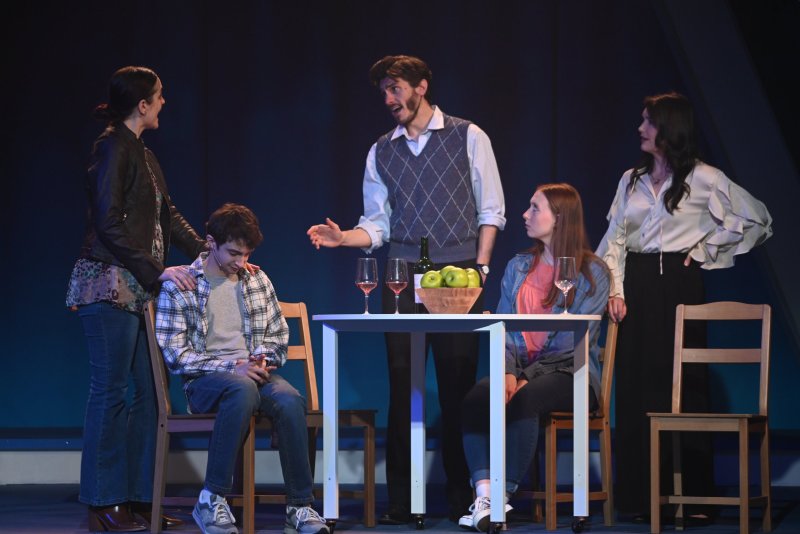 Five actors on stage; a young man sits at a table with a woman, while two men and another woman stand around them.
