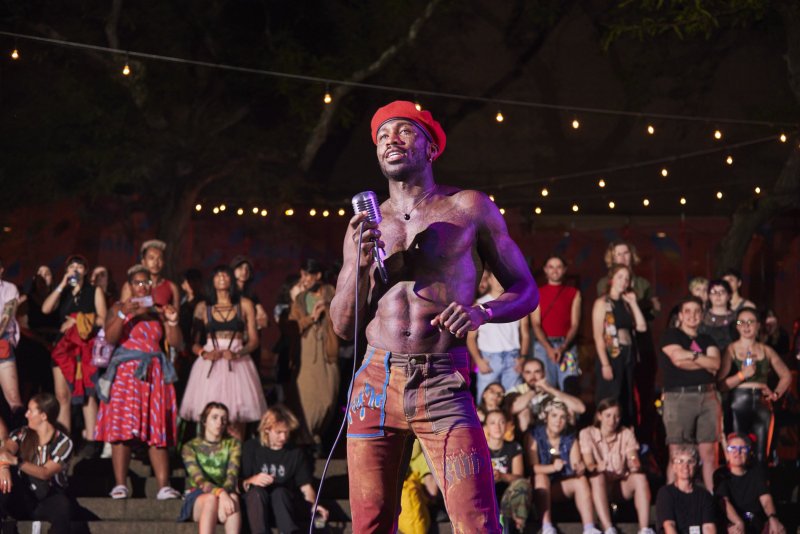 Performance view: shirtless Black person in red beret sings into a mic before a crowd. String lights overhead, dark background. Person wears patterned pants.