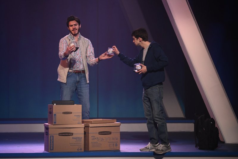 Two men on a stage, one in a plaid shirt and vest, the other in a blue hoodie, exchanging small objects over cardboard boxes.