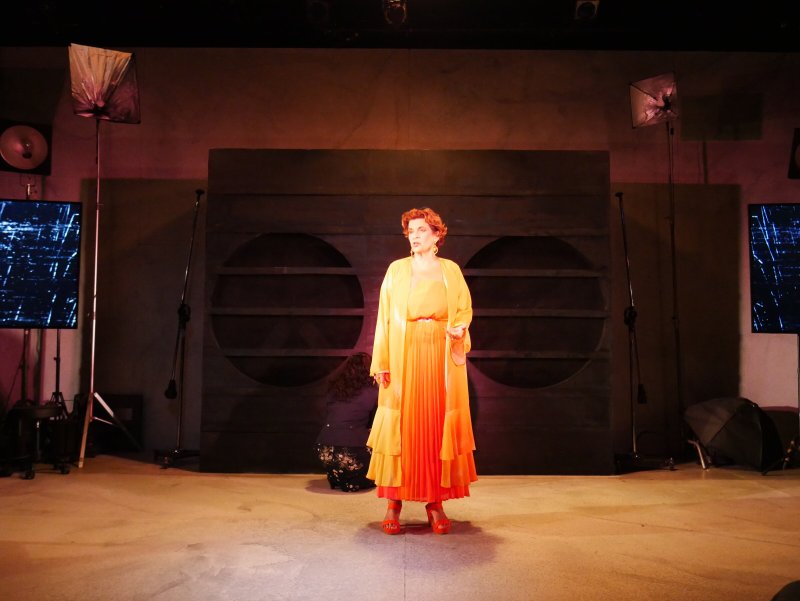Woman in flowing orange dress and coat on stage, lit by warm light, with dark panels and screens in background.