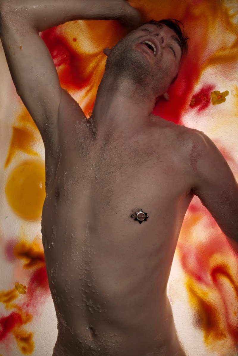 Bare-chested man with nipple piercing, head back, mouth open, against a background of red, orange, and yellow liquid.