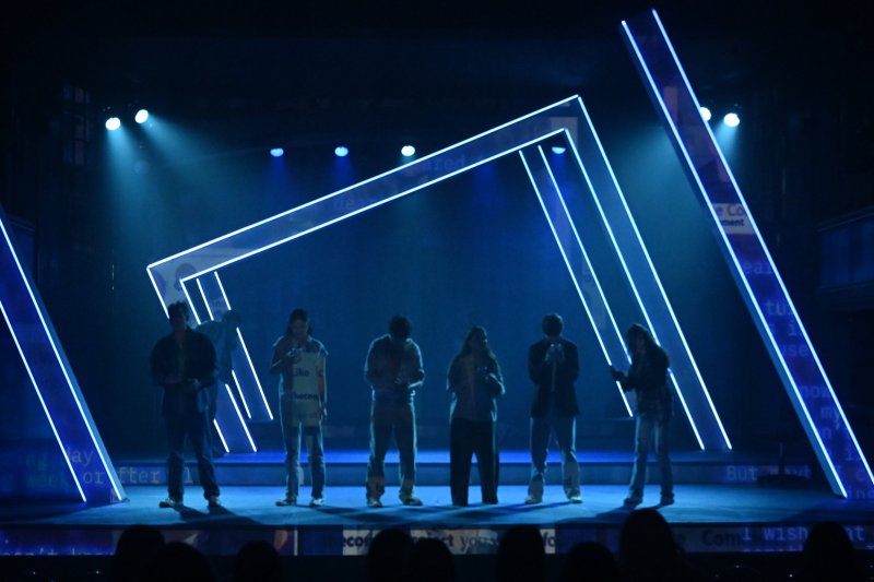 Six figures stand on a dark stage with glowing blue geometric frames and projected text, illuminated by spotlights.