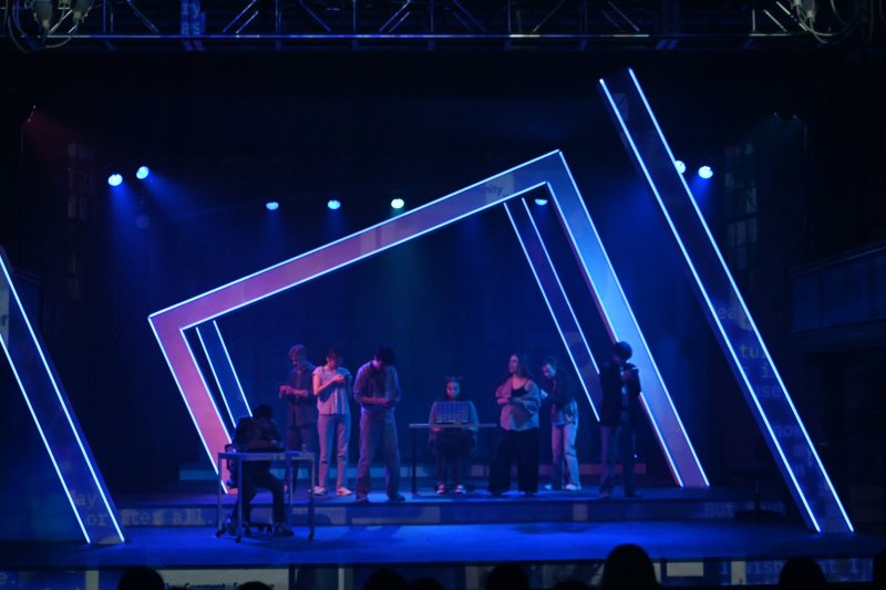 Stage production of Dear Evan Hansen with actors in casual clothes under blue lights, framed by glowing white and blue geometric outlines.