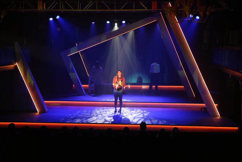 Stage with a central performer in a spotlight, flanked by two figures in shadow and geometric orange-lit frames.