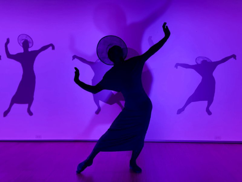 Silhouette of a person in a wide-brimmed hat and dress dancing, with two fainter silhouettes behind, all against a purple wall.