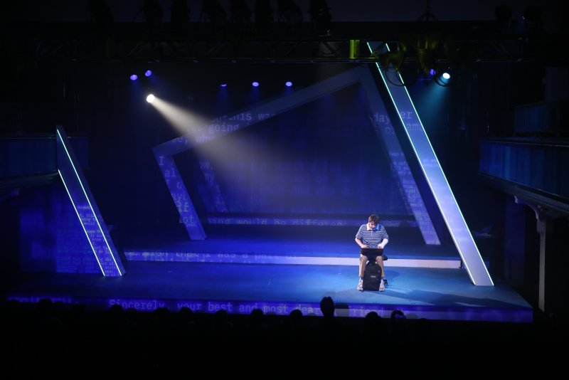Stage production with a lone male actor sitting center, bathed in a bright spotlight against a dark blue background with projected text.