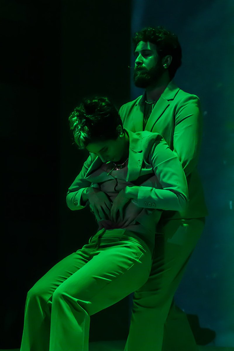 Two performers in green suits, one standing behind the other who is seated, under green stage lighting.