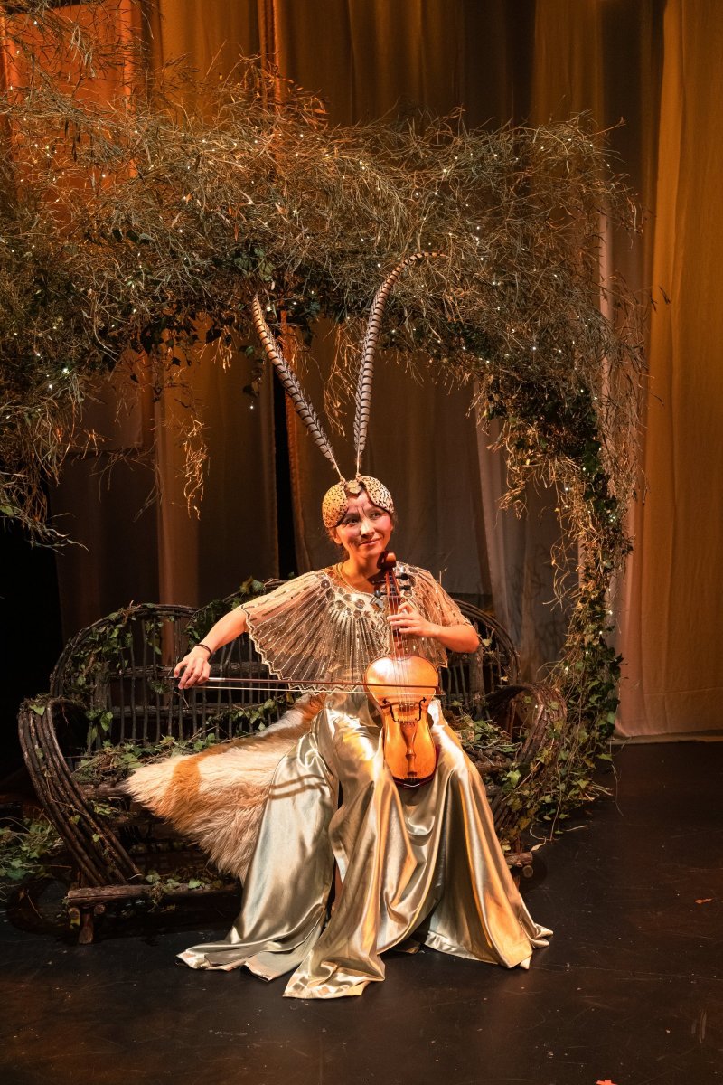 Woman in gold dress and feathered headpiece playing violin on a rustic bench under a leafy arch with twinkling lights.
