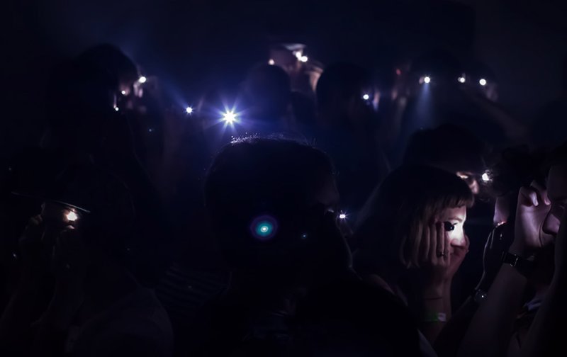 Dark photo of a crowd with faces illuminated by bright phone flashlights, creating lens flares and starbursts.