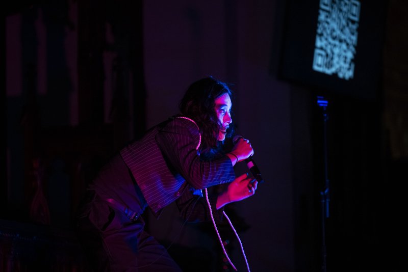 Performer with long dark hair, wearing a pinstripe jacket and pearl necklace, singing into a microphone under blue and red stage lights.