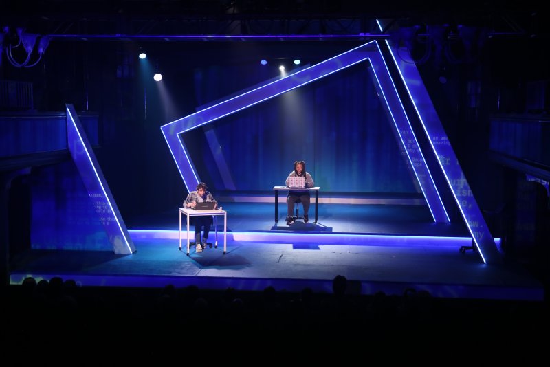 Two actors on a dark stage, illuminated by blue light, sit at desks with laptops, framed by glowing blue geometric structures.
