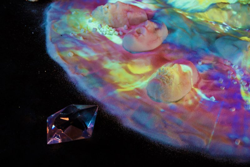 Close-up of a dark surface with a faceted crystal and a colorful projection illuminating white rocks and granules.