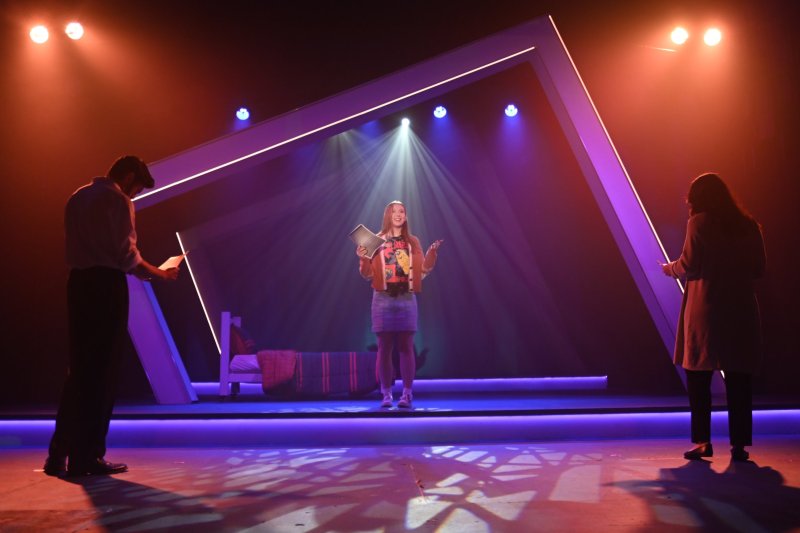 Three actors on a stage with a minimalist set, bathed in dramatic purple and orange lighting.