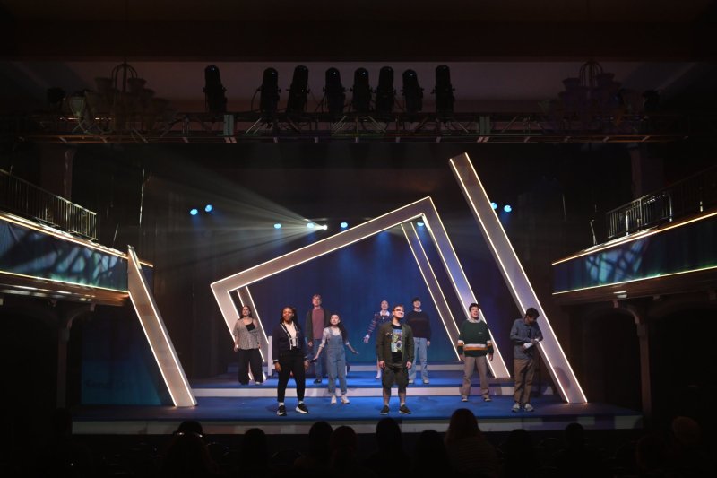 A stage production with ten actors, some standing, some walking, amidst large, glowing white geometric frames.