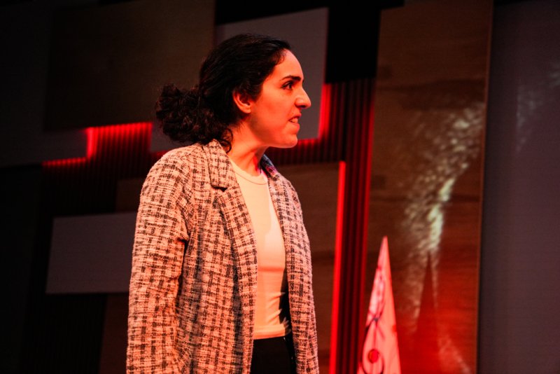 Woman with dark curly hair in a plaid blazer and white shirt, looking right with a tense expression, red and dark background.