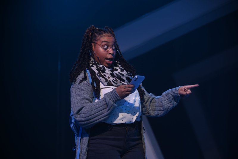 Woman with braids, glasses, and a patterned scarf, holding a phone and pointing right, against a dark blue background.