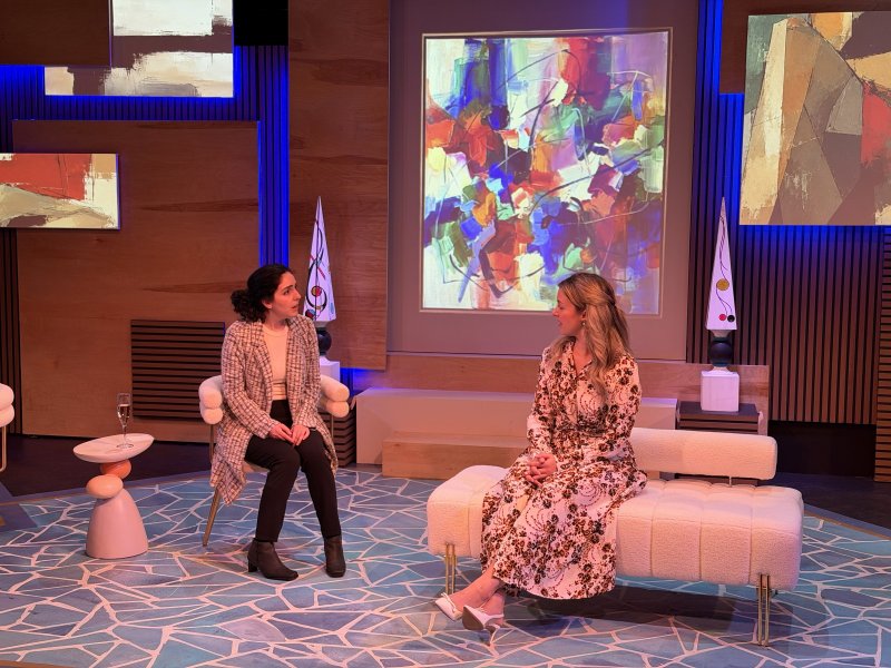 Two women converse on a stage set with abstract art projections, modern furniture, and blue geometric floor patterns.