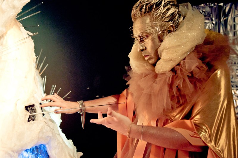 Performer in gold paint and costume with ruffled collar, interacting with a white, spiky sculpture glowing blue.