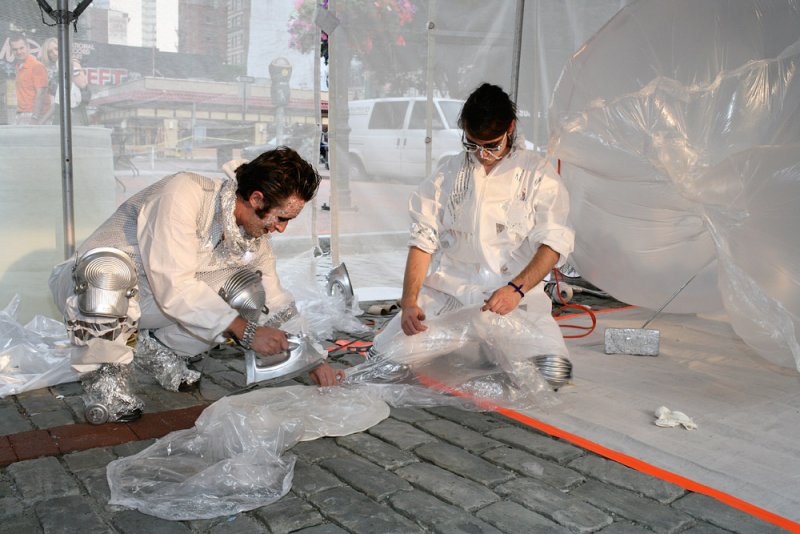Two performers in white and silver costumes iron clear plastic on a brick ground, with a city street in the background.