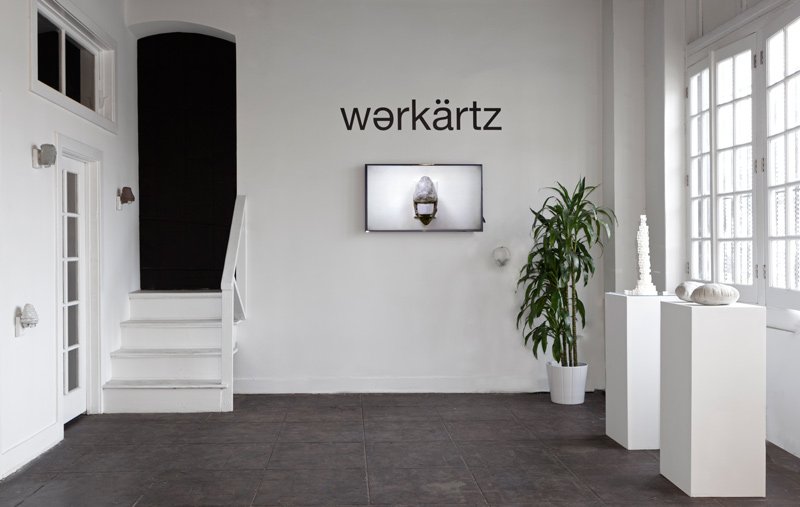 Gallery interior with "wərkärtz" on white wall above a TV displaying a textured object; stairs, plant, and sculptures flank it.
