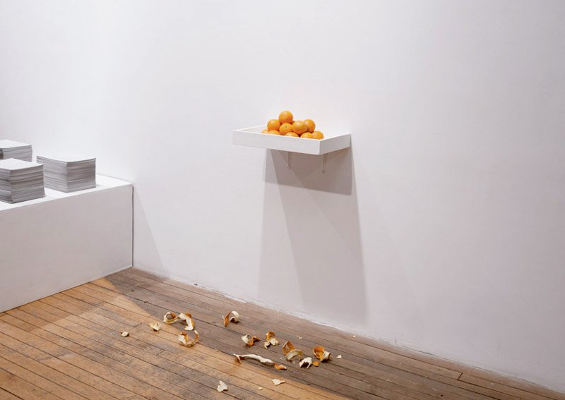 White shelf with oranges on a white wall, with orange peels scattered on a wooden floor.