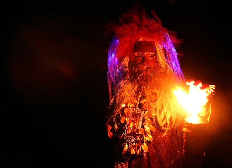 Person in a dark mask with a long beard, wearing a large necklace, holding a torch with a bright flame.