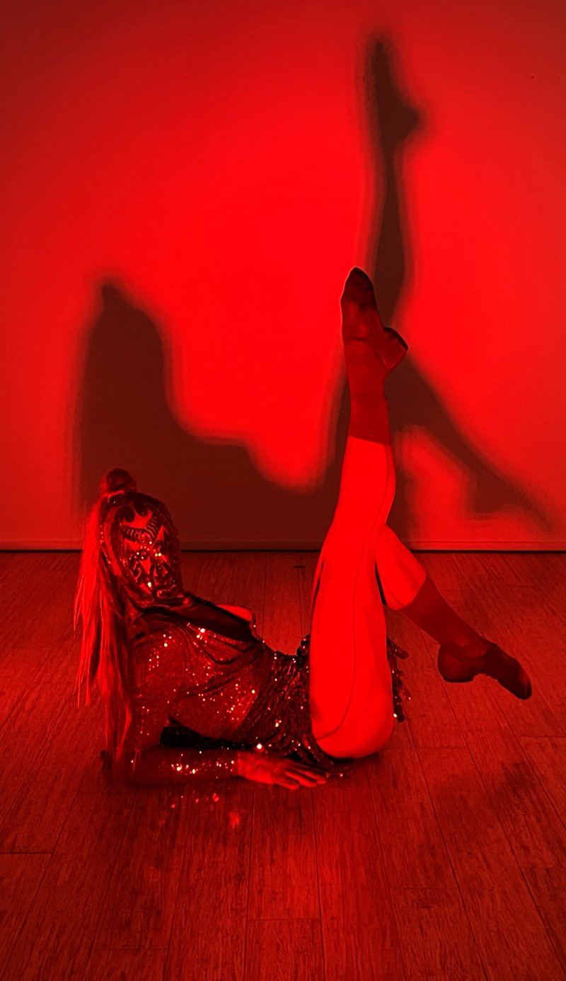 Performer in a sequined costume lies on a wooden floor, one leg raised, casting dramatic shadows on a vibrant red wall.