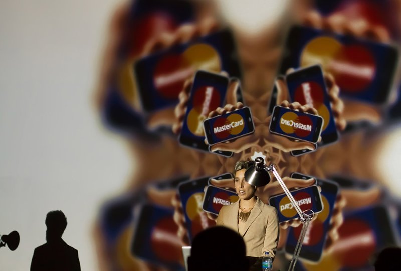 Woman speaking at a podium with a repeating pattern of hands holding MasterCards projected behind her.