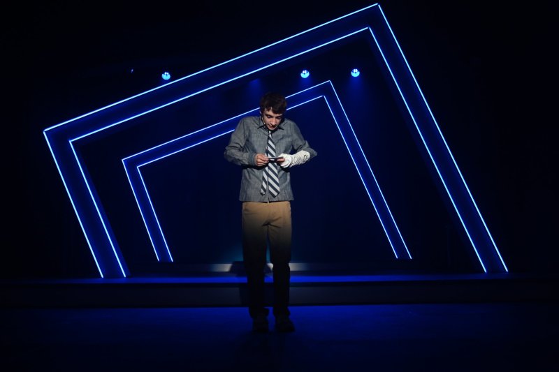 A young man in a gray shirt and striped tie, with a cast on his arm, stands on a dark stage with blue neon geometric outlines.