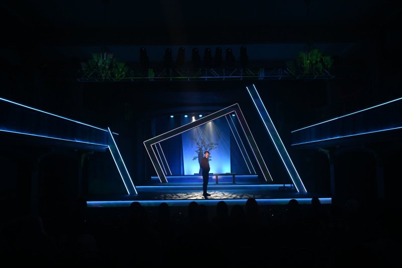 Stage performance with a lone figure holding a plant, illuminated by a spotlight and surrounded by glowing blue geometric lines.