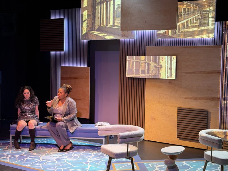 Two women on a stage with a backdrop of wooden panels and projected images, one seated and one gesturing while holding a book.