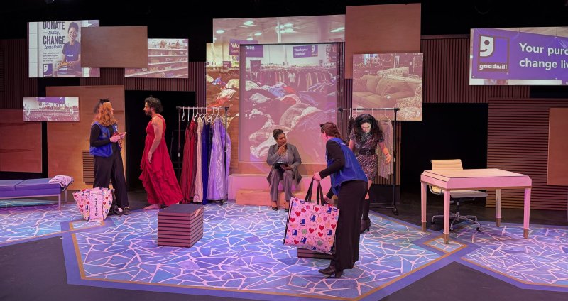 Stage production with five actors, a clothing rack, and projected images of a Goodwill store on wooden panels.