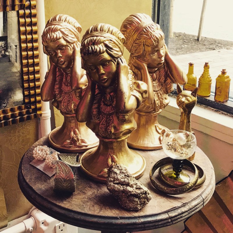 Three golden busts of women with hands to their ears on a wooden table, with a golden cat figurine and bottles.