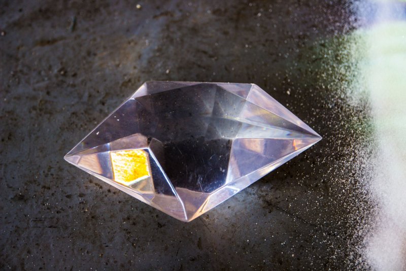 Clear, faceted crystal gem with a yellow reflection, centered on a dark, speckled surface.