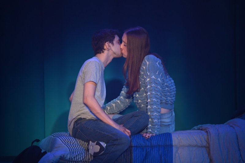 A young man and woman kiss on a bed, bathed in blue light. He wears a gray shirt and jeans; she wears a blue sweater.
