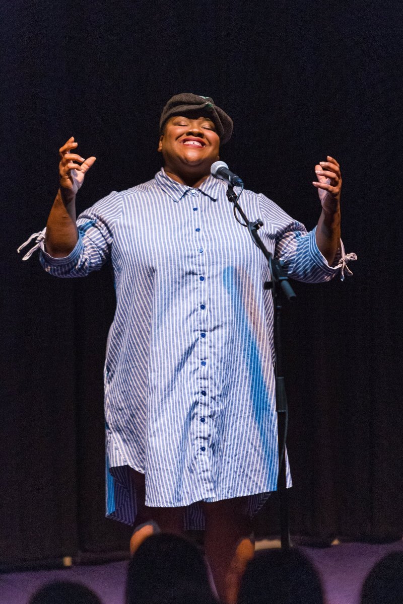 Performer on stage, lit from above, smiles with hands raised. Wearing a blue and white striped shirt dress and a newsboy cap. Microphone stand in front of a dark background.
