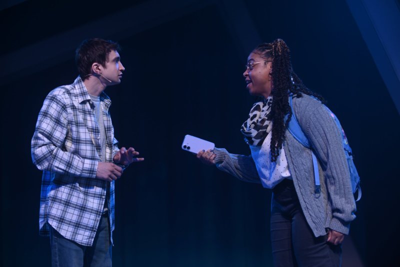Two actors on a dark stage, illuminated by blue light. A man in a plaid shirt gestures, facing a woman in glasses holding a phone.