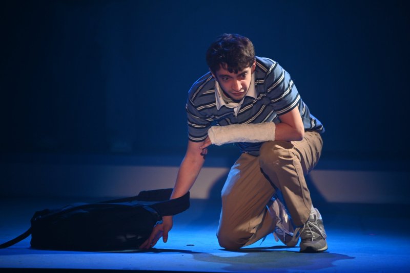 A young man with a cast on his arm kneels on a dark stage, looking forward with a concerned expression.