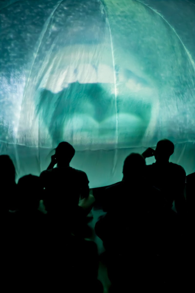 Silhouetted figures watch a large, translucent dome illuminated with swirling teal and white light.