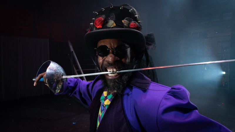 Man in purple coat, spiked hat, and sunglasses holds a sword in his mouth. Stage lighting.