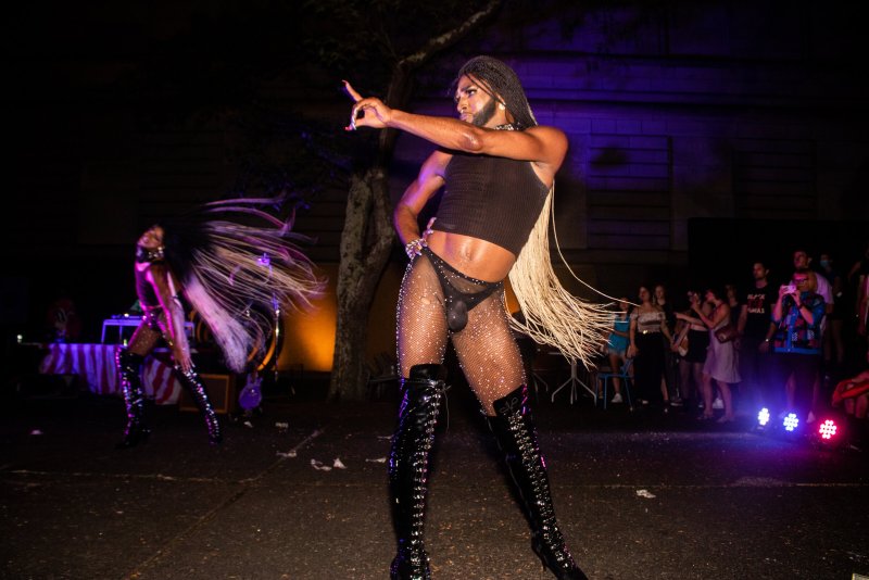 Drag performer in a black tank top, fishnet stockings, and knee-high boots poses on stage under purple lights.