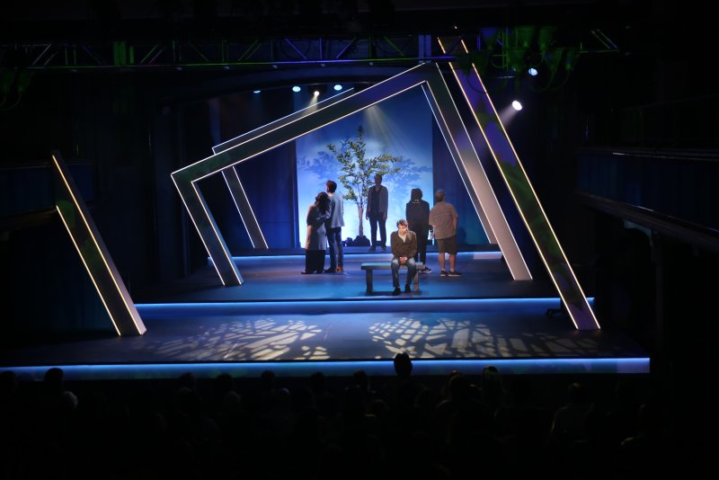 A dark stage with glowing white geometric frames, a tree, and actors. A lone actor sits center, others stand around.