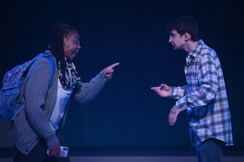 Two actors on a dark stage. A woman in glasses and a backpack points at a man in a plaid shirt, who gestures back.