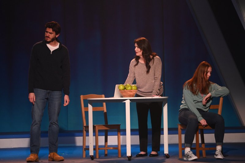 Three actors on stage: a man standing left, a woman standing center by a table with green apples, and a woman seated right.