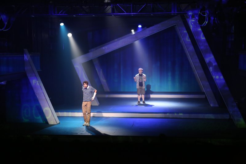 Two young men on a dark stage with blue lighting and geometric set pieces. One man wears a striped shirt, the other a t-shirt and shorts.