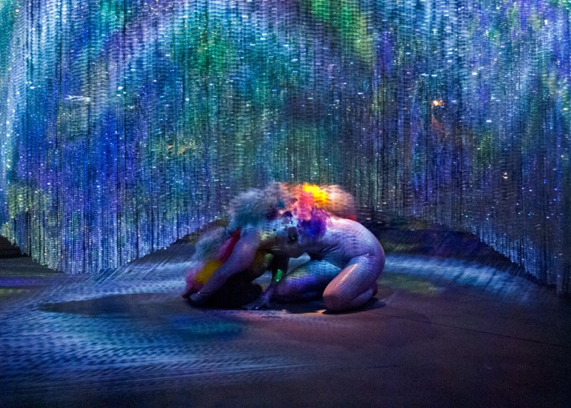 Performance art with a figure kneeling on a dark stage, surrounded by shimmering blue, green, and purple light curtains.
