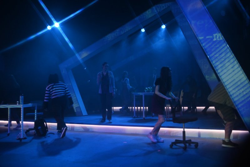 Stage bathed in blue light, performers in casual clothes, one standing center, others moving around tables and chairs.