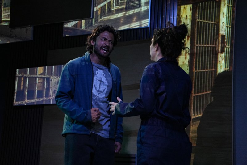 Two actors on stage, a man in a blue jacket yelling at a woman in a dark jumpsuit, with projected images of prison cells behind them.